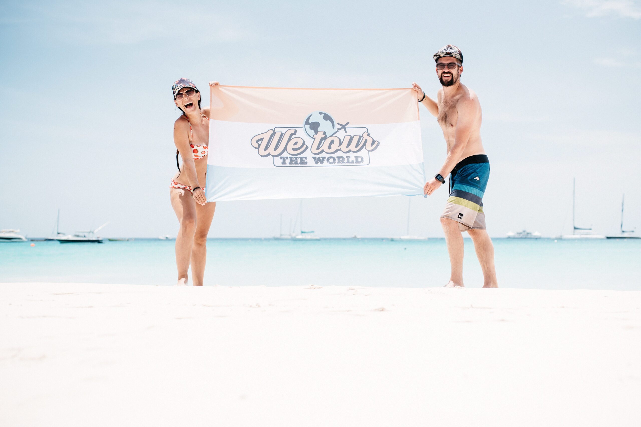Bianca and Tom holding the We Tour The World flag on a white Caribbean beach
