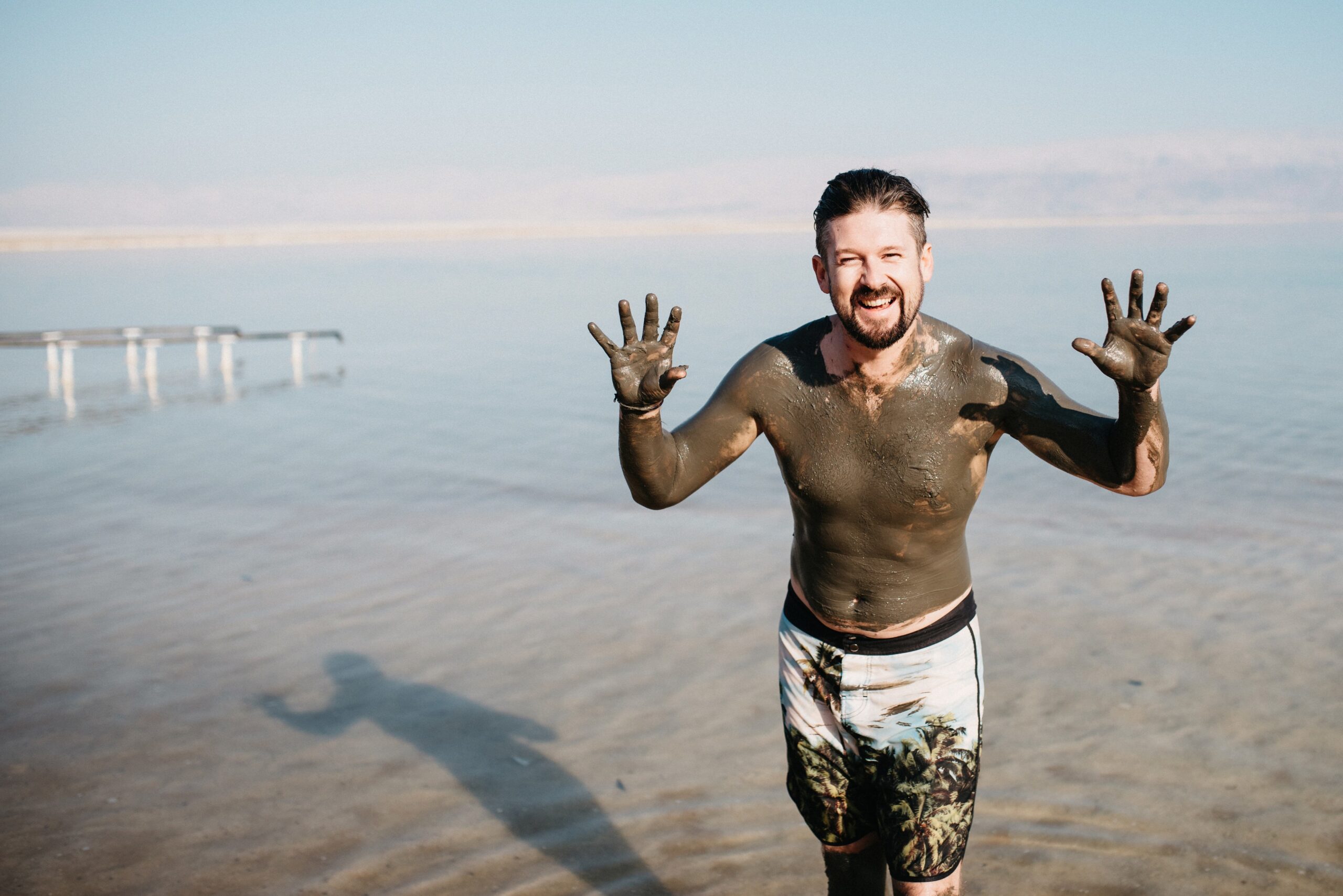 Tom standing in the Dead Sea covered in dark mineral mud with both hands raised